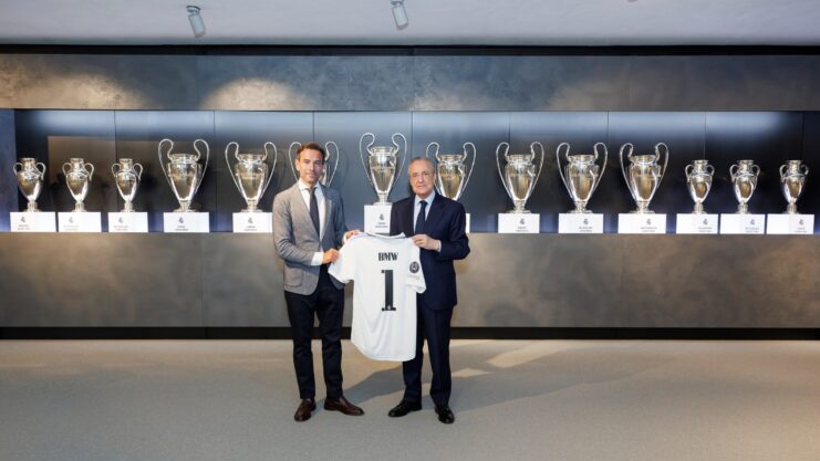 Florentino Pérez e Manuel Terroba segurando camisa do Real Madrid em sala com troféus do time em estante atrás.