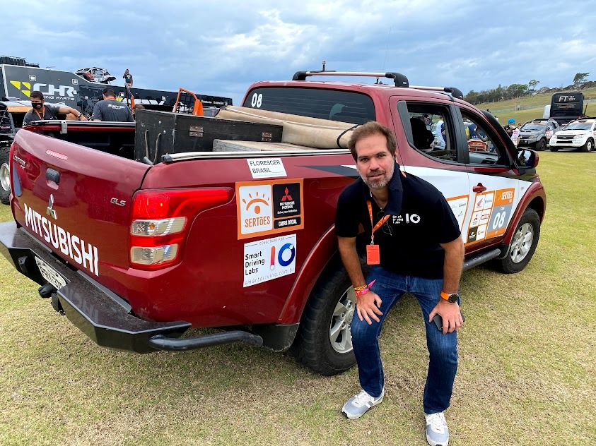 Homem posa para foto ao lado de um carro Mitsubishi L200 Triton. O carro é vermelho e possui adesivos de patrocinadores: Rally dos Sertões, Mitsubishi Motors, Smart Driving Labs, Florescer Brasil.