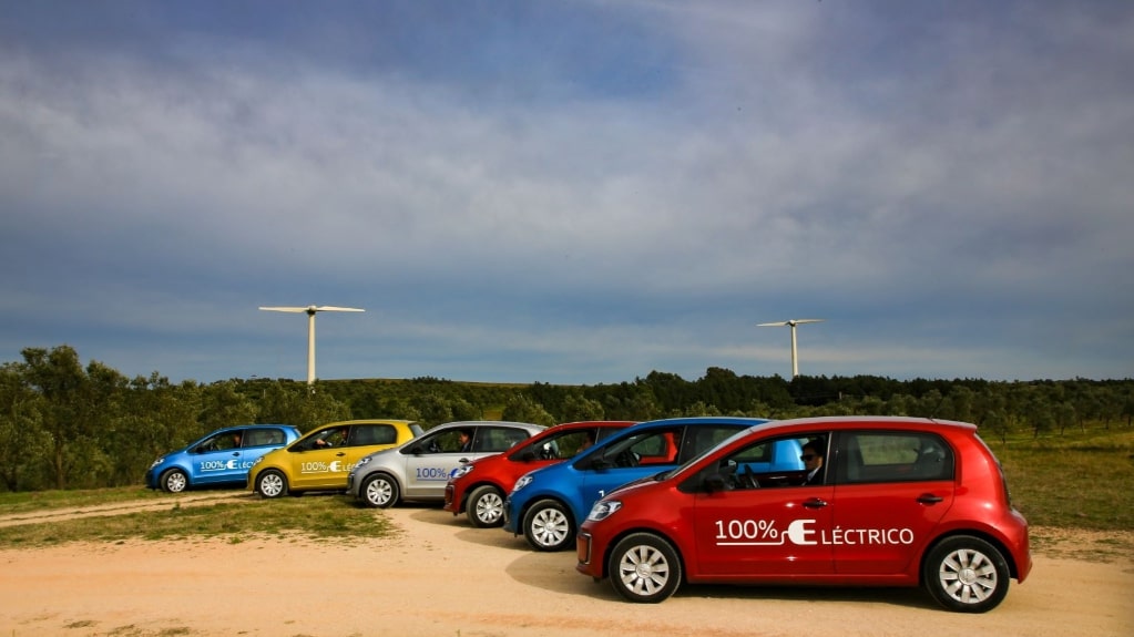 Unidades do VW e-up! estacionadas lado a lado em área verde ao ar livre, com chão de terra. Eles são de cores azul, amarela, cinza e vermelha 