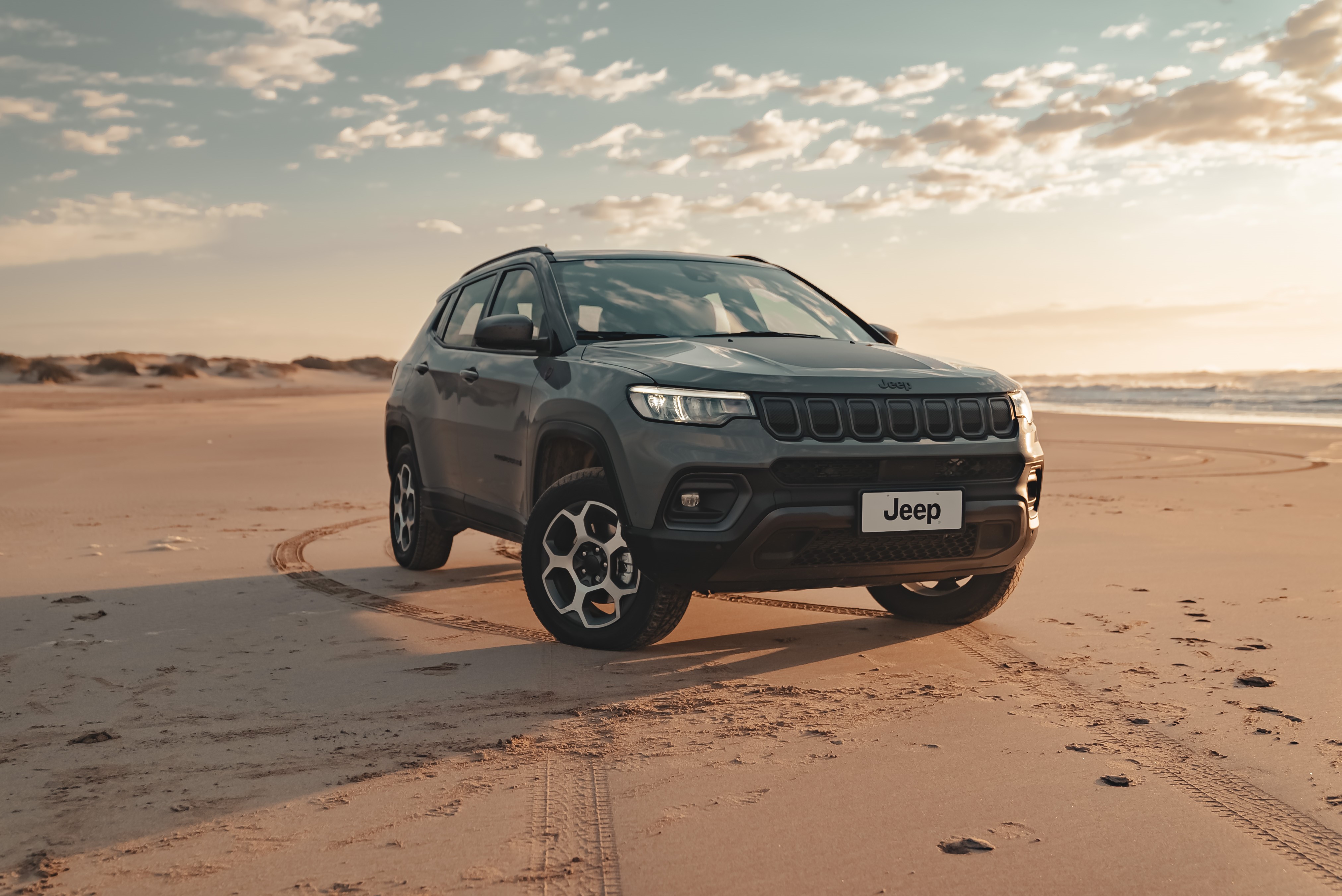 Jeep Compass em tom acinzentado visto da diagonal frontal, em meio à uma praia e sobre a areia. 