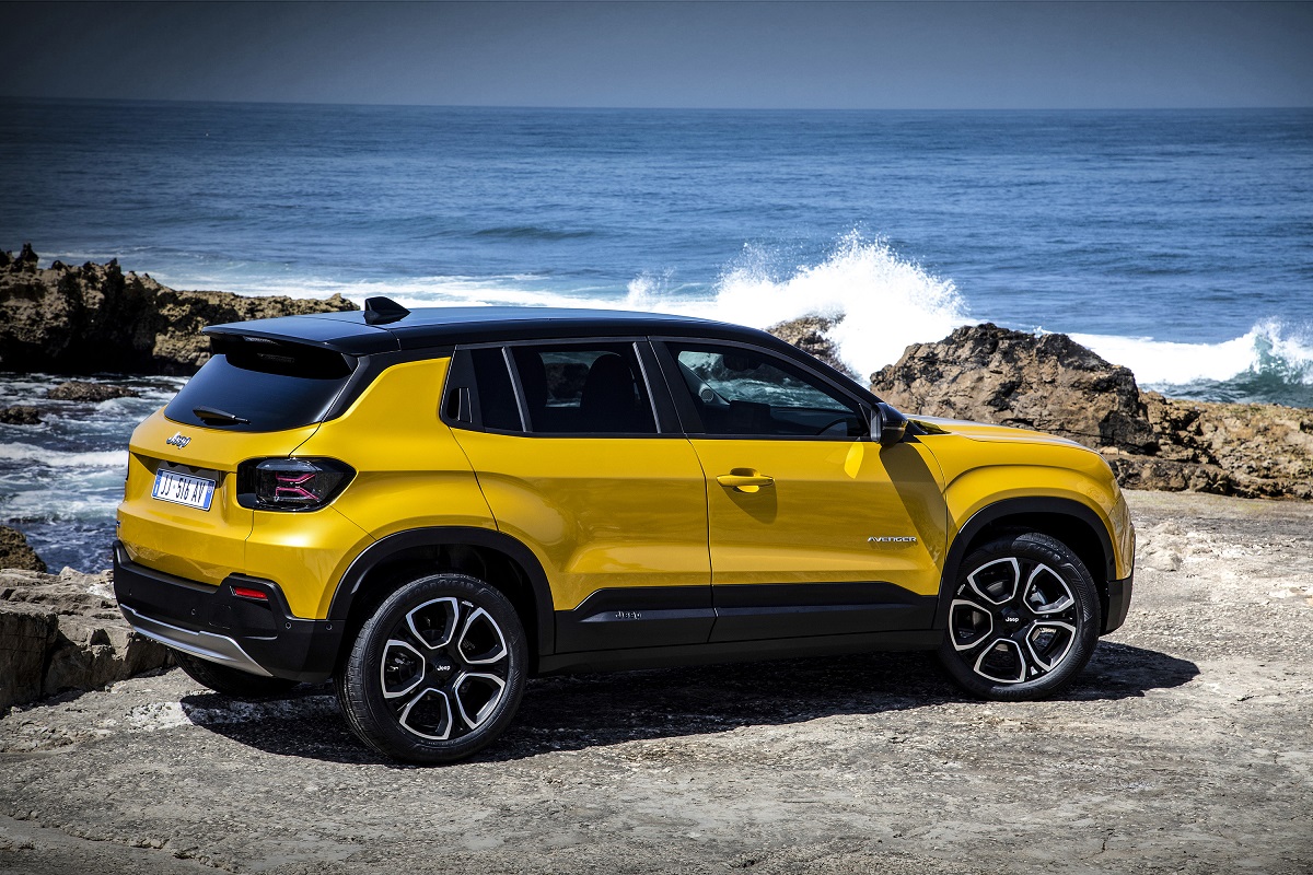 Jeep Avenger amarelo visto da diagonal traseira. Ele está posicional ao lado de rochas e mar. 