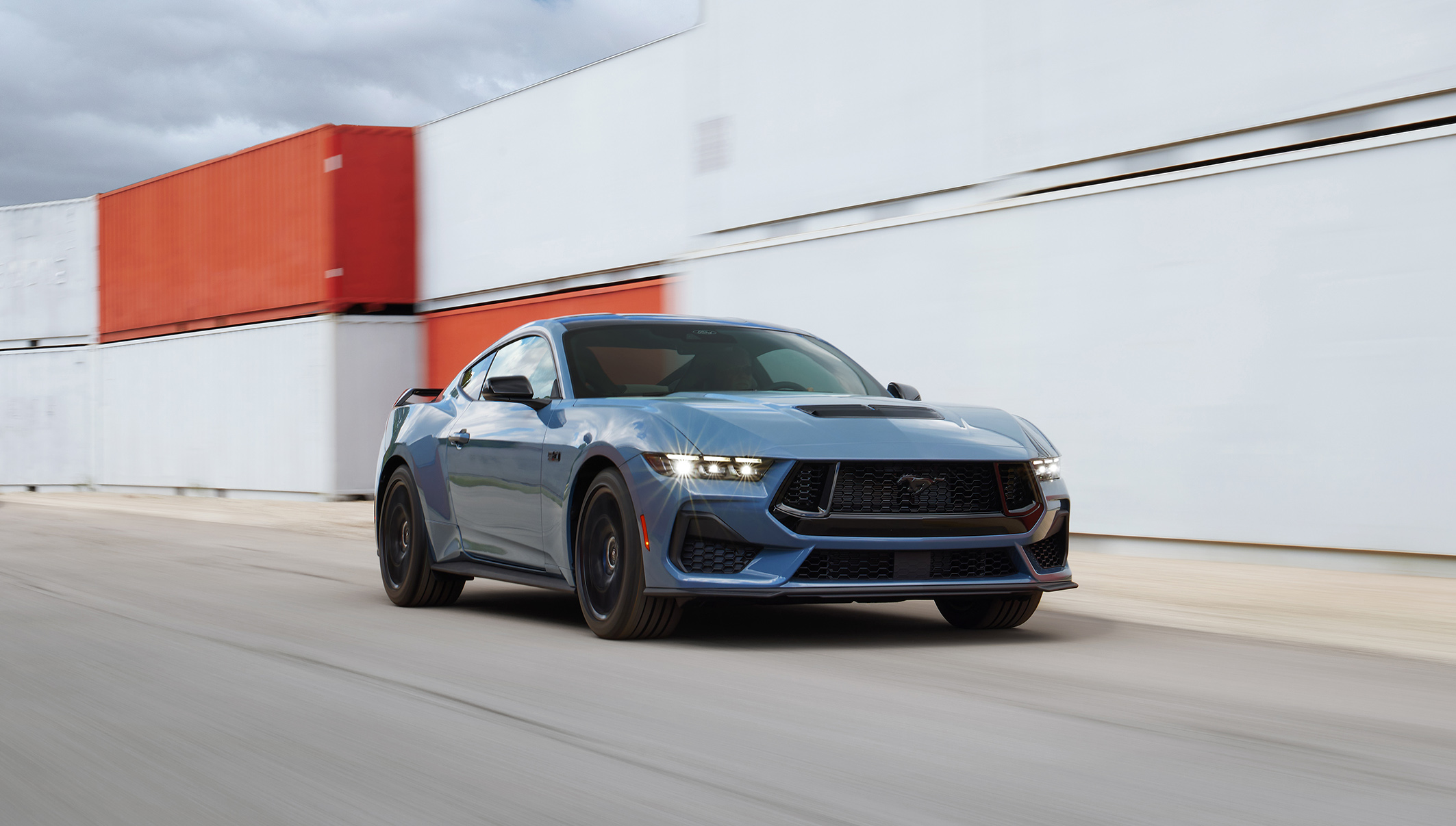 New seventh generation Mustang in blue tone, viewed from the front diagonal, moving in a multi-container space. 