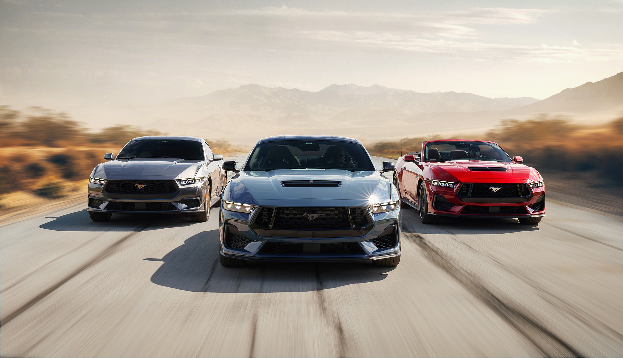 There are three 2024 Ford Mustang models from the seventh generation.  A blue model, a red model and a gray model, all seen from the front moving down the road.