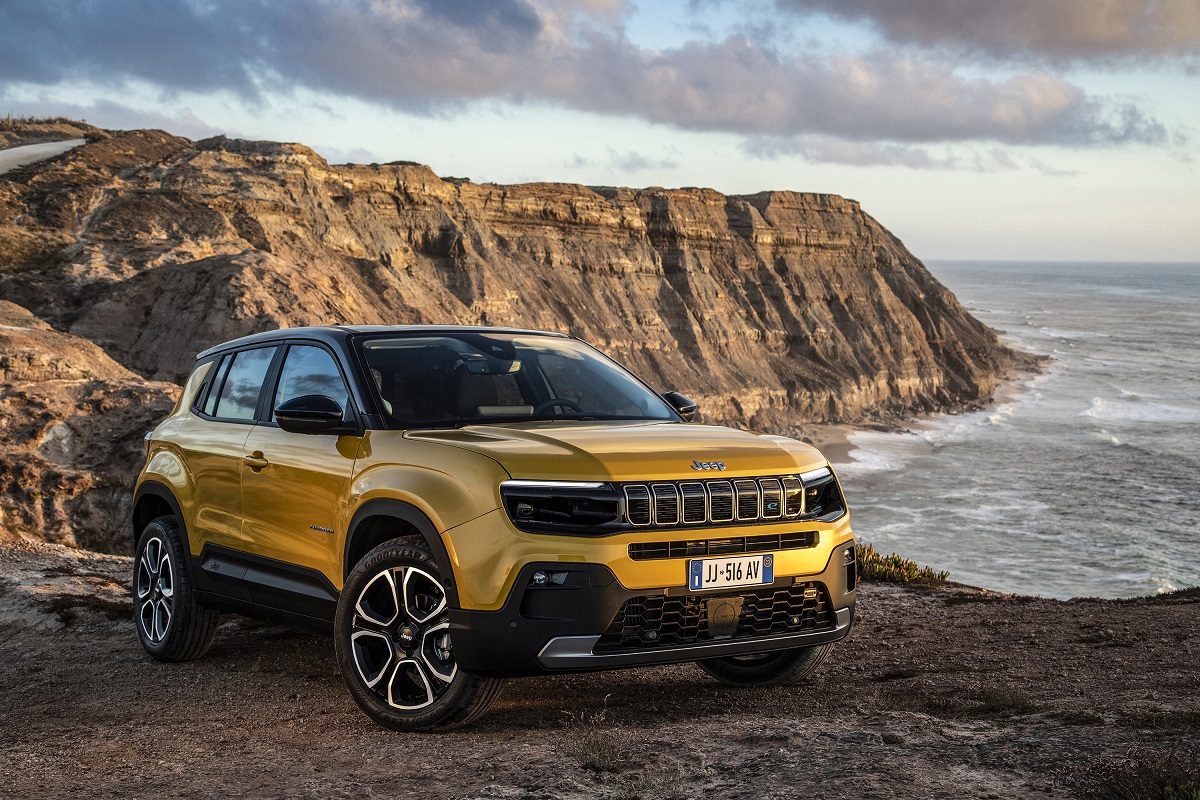 Jeep Avenger amarelo visto da diagonal frontal. Ele está posicional ao lado de penhasco com mar e montanhas. 