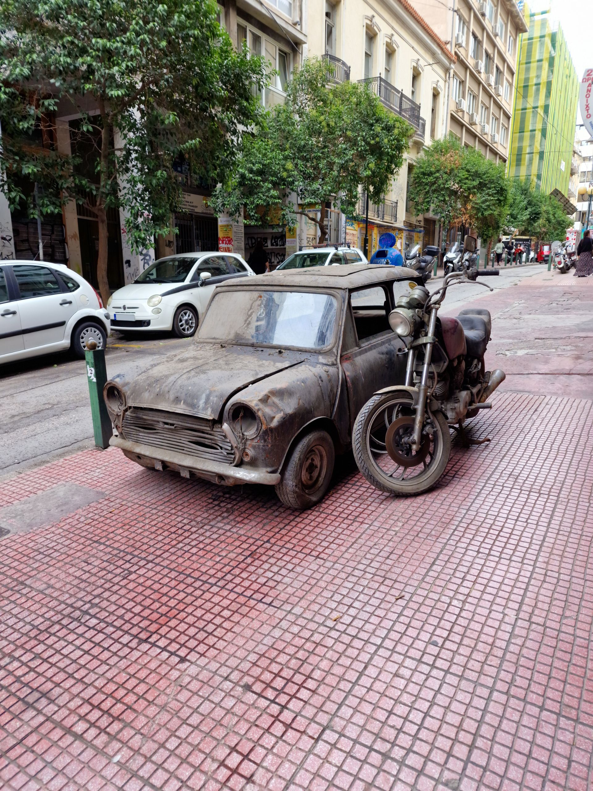 Mini Cooper e moto Yamaha em péssimo estado estacionados na calçada. Eles estão cobertos de sujeira e ferrugem.