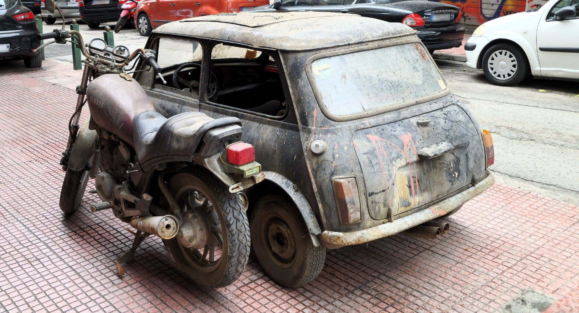 Mini Cooper e moto Yamaha completamente deteriorados vistos da diagonal traseira e estacionados na calçada.