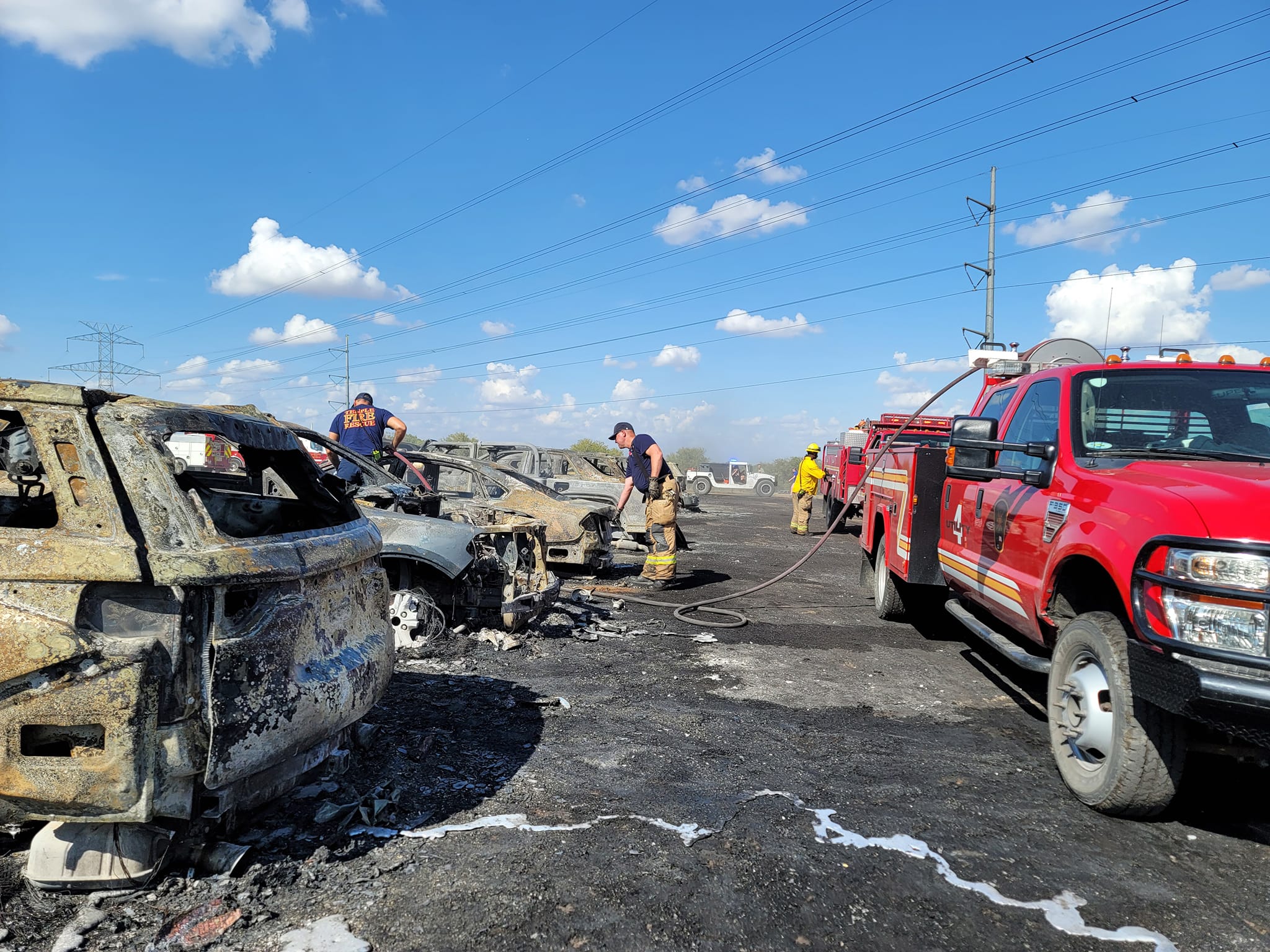 Visão do estacionamento da fazenda com vários carros destruídos e queimados. Há bombeiros trabalhando para apagar o fogo. 