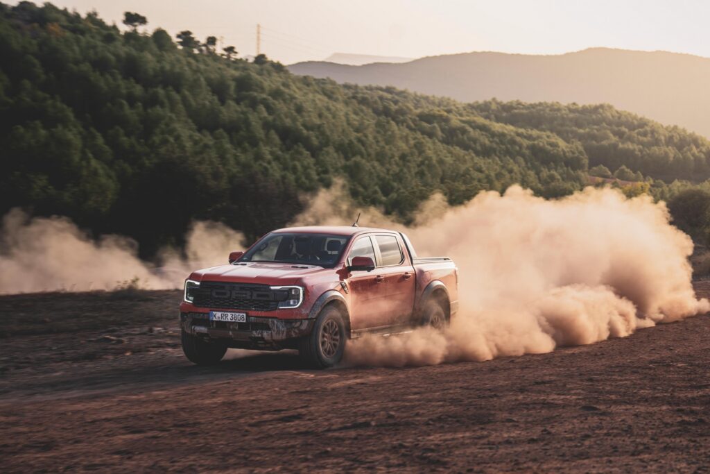 Ford Ranger Raptor laranja vista da diagonal frontal andando em chão de terra. Há muita poeira sendo produzida pelos pneus.