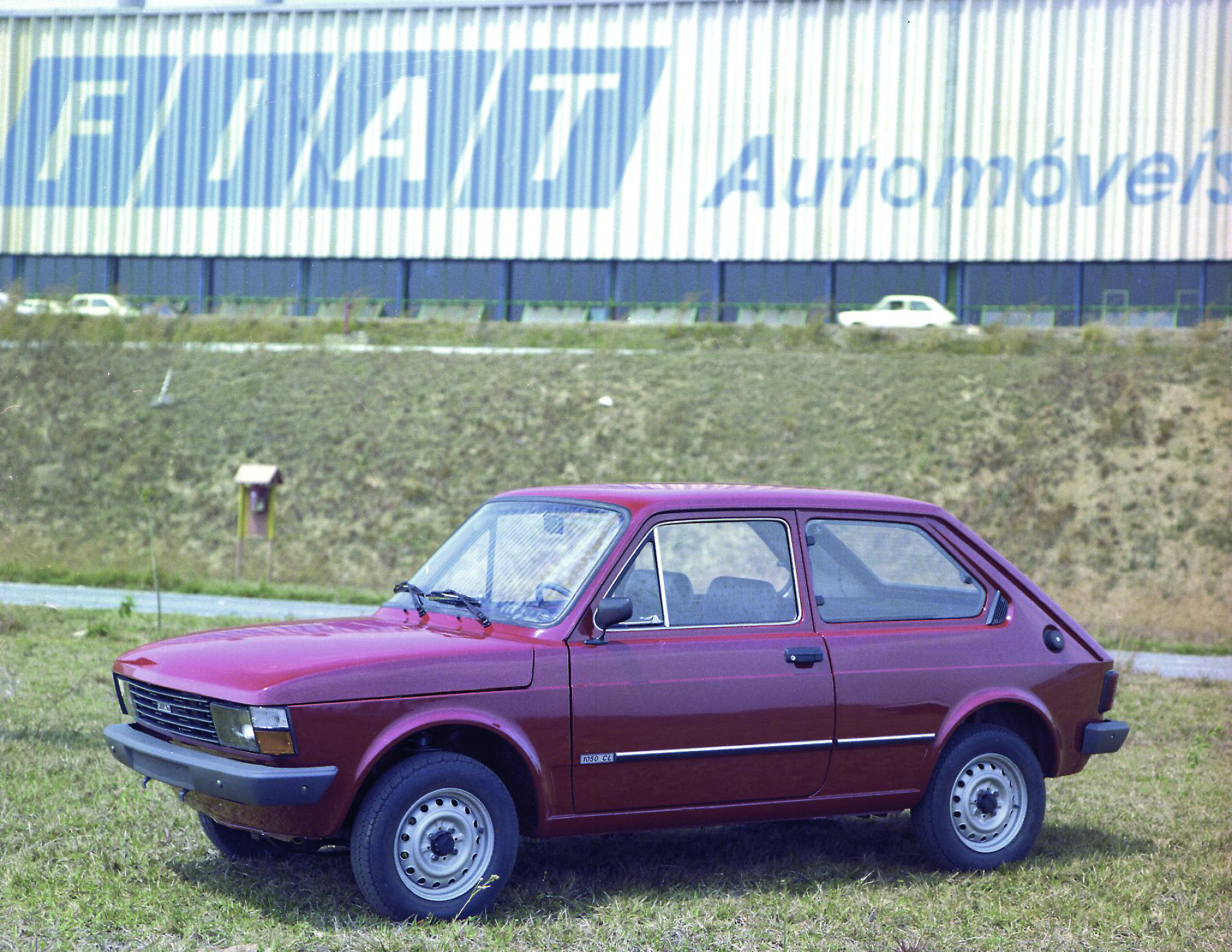 Fiat 147 GL vermelho de frente com fábrica de Betim (MG) ao fundo