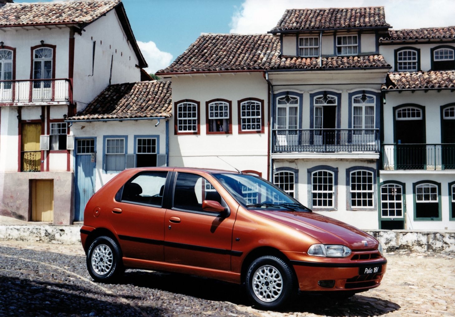 Fiat Palio 16V 1996 vermelho visto a estacionado partir da lateral com casario colonial ao fundo