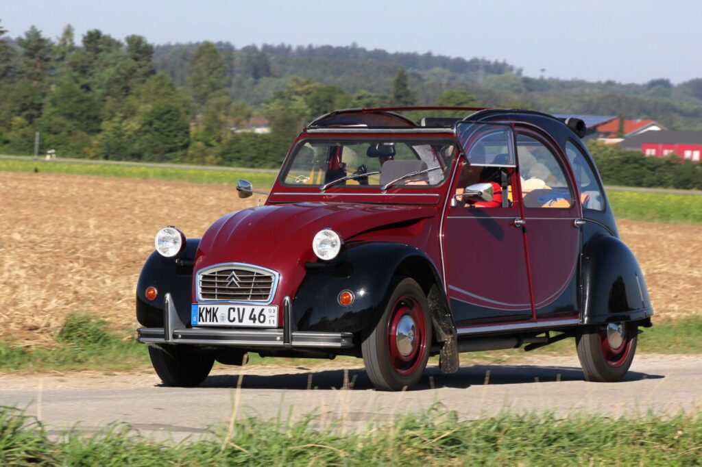 Renault 2CV em tom vinho e preto visto da diagonal frontal em movimento na estrada.