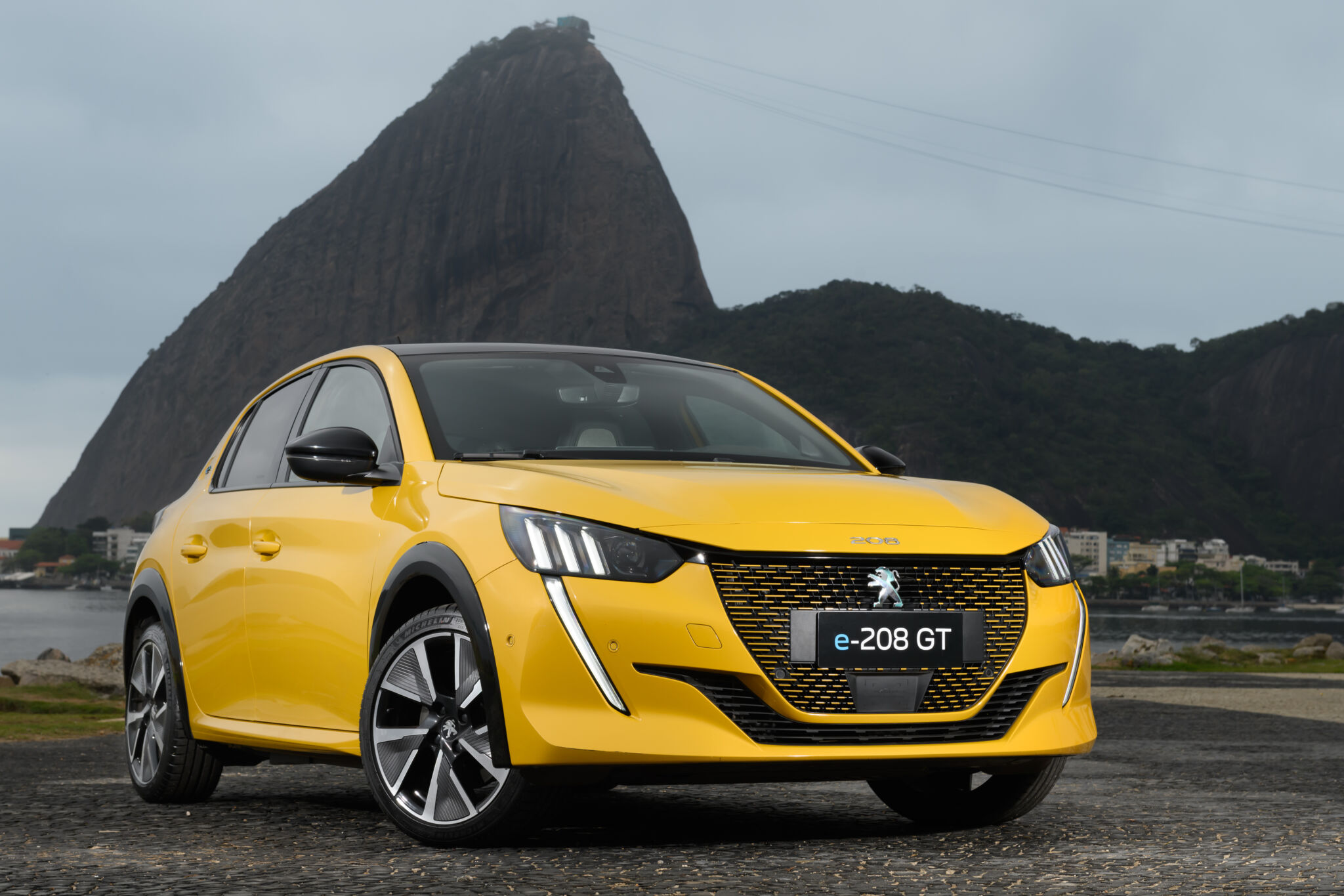 Peugeot e208 GT amarelo de frente estacionado com morro do Pão de Açúcar ao fundo;; para matéria dos carros elétricos mais baratos do Brasil.