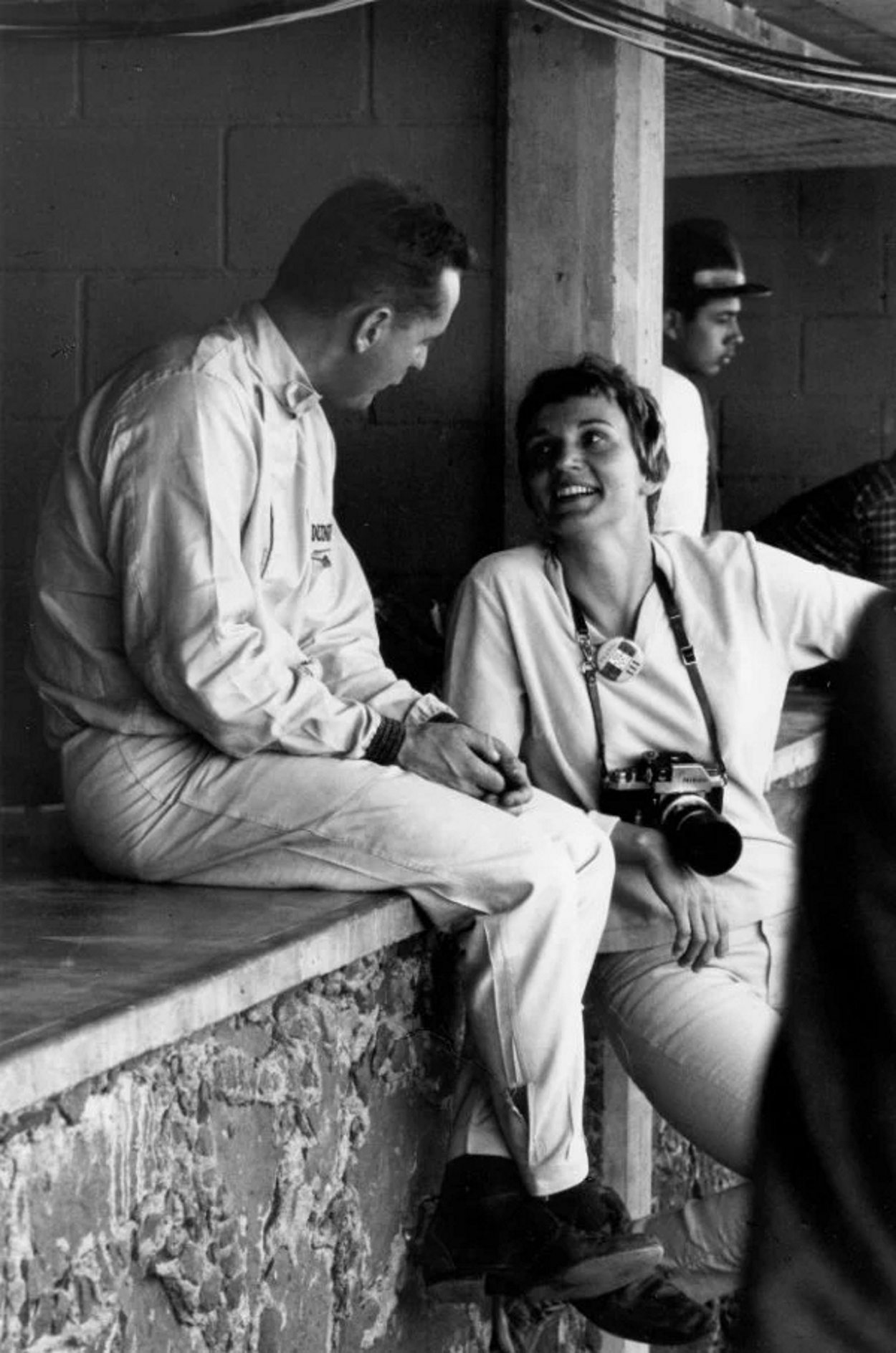 Denise McCluggage com uma câmera fotográfica ao lado de um piloto. Ela é homenageada no Dia da Mulher.