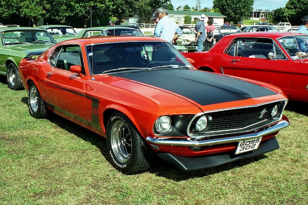 Ferrari Mustang Boss 302 vermelho em área de grama junto de outros veículos.  O modelo é um dos que serão transformados em carros elétricos pelo ator de Homem de Ferro. 