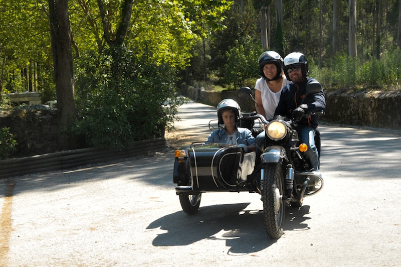 Um motorista sobre uma moto acoplada a um side car com um turista atrás dele e um a bordo do mini carro, todos usam capacete.