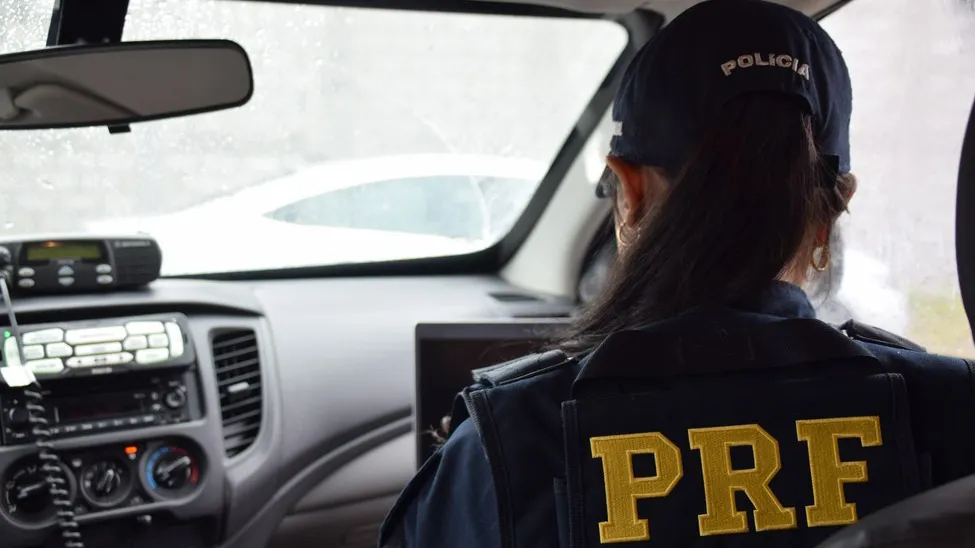 Mulher vista de costas dentro de banco da frente de carro vestida com uniforme e boné da PRF